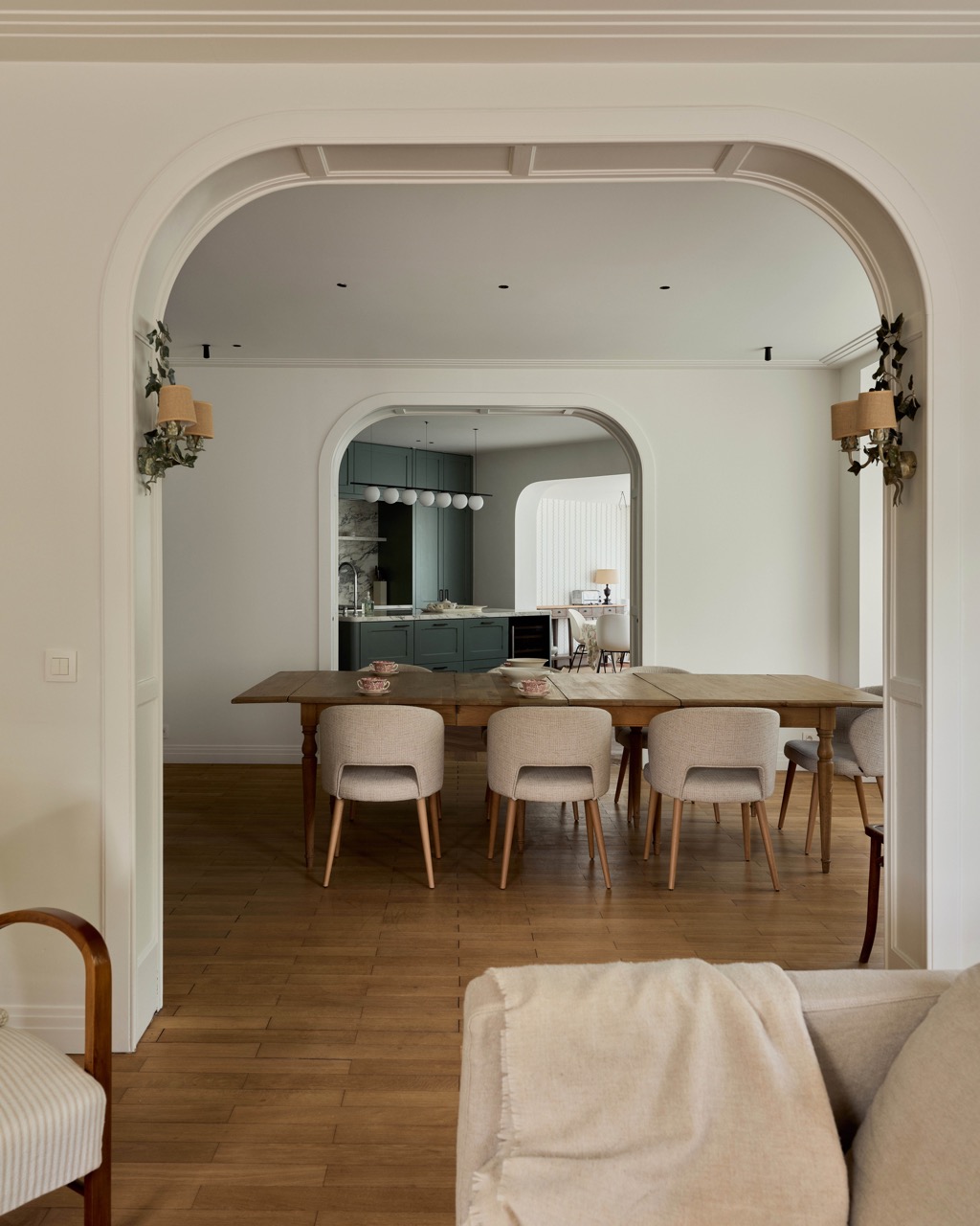 Salle à manger avec table en bois et chaises beiges, vue sur une cuisine verte derrière une arche.