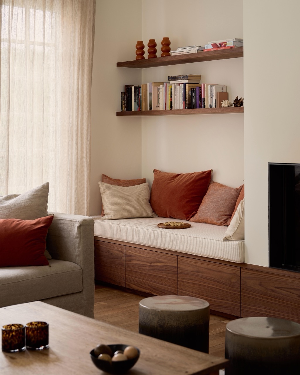 Banc de lecture avec coussins orangés, étagères garnies de livres et vases, fauteuil gris et table basse en bois